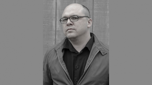 Matthew Olzmann in gray scale against a wood exterior. He wears glasses and his chin is titled up.