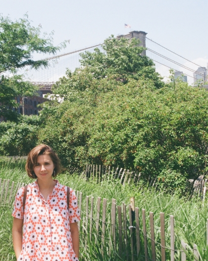 Kate stands in front of some trees in Brooklyn Bridge park. Behind her is lots of green, and the bridge.